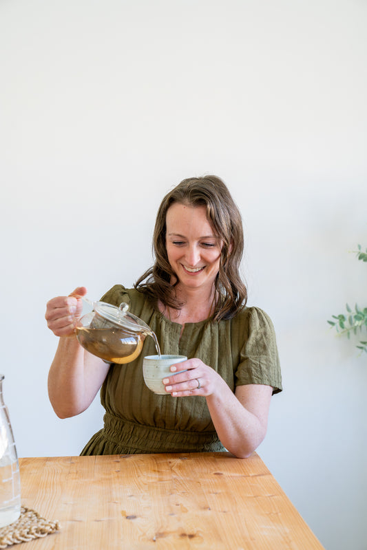 Woman pouring tea from a glass jar into a small cup on a wooden table. Order custom tea blend or organic herbs by filling out the contact us form. 
